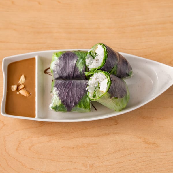 Four fresh spring rolls with purple shiso leaves and a side of peanut sauce with crushed peanuts.
