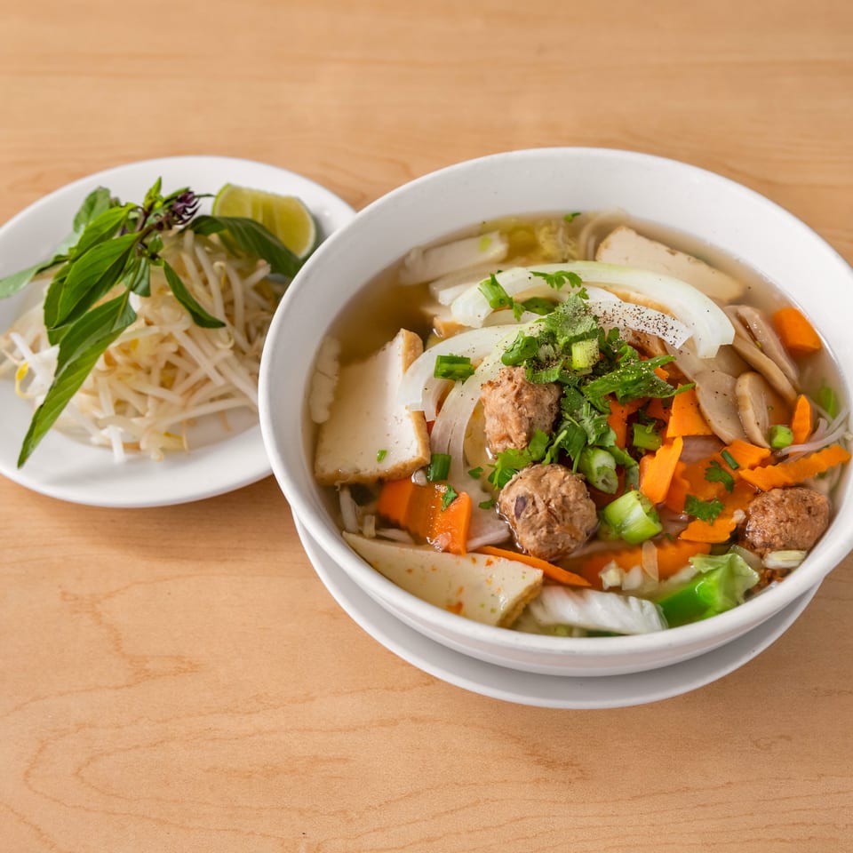 A bowl of Vietnamese pho with meatballs, vegetables, and a side plate of bean sprouts and basil.