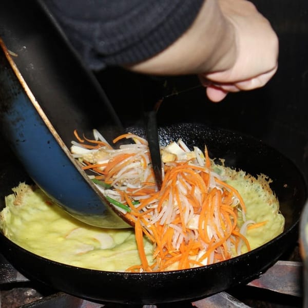 A chef's hand adds shredded carrots and bean sprouts to a vegan omelet in a pan.